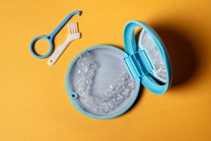 Clear aligner trays in an open blue storage case with a mirror, beside an aligner removal tool and a small cleaning brush on an orange background
